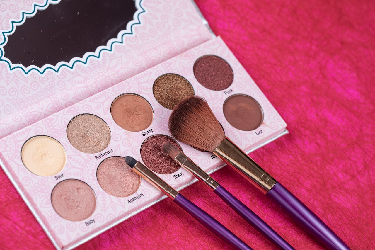 Services A detailed close-up of an eye shadow palette with makeup brushes on a vibrant pink fabric background.
