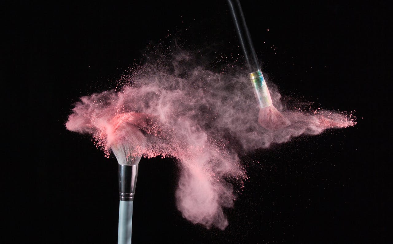 Services Close-up of makeup brushes colliding, releasing a cloud of pink powder against a black background.